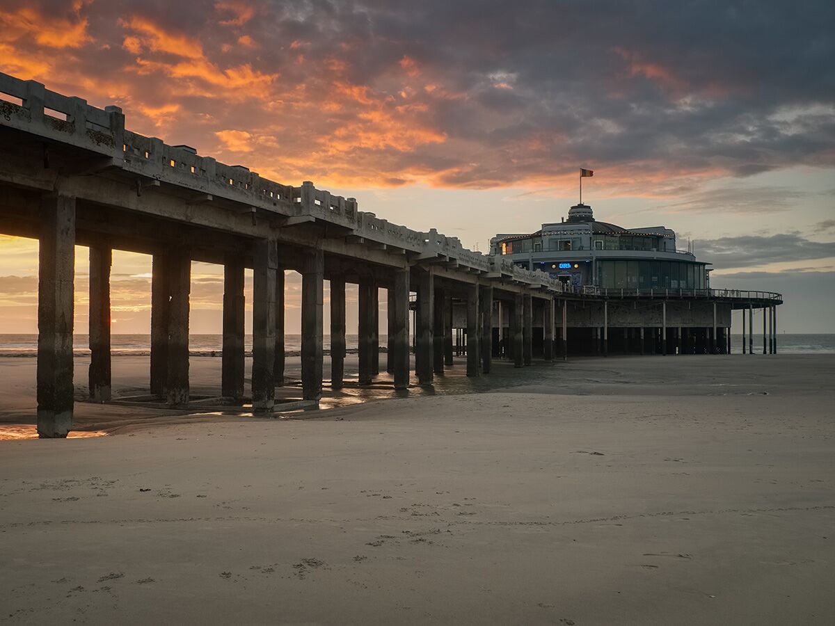 Lange Seebrücke bei Sonnenuntergang über Sandstrand mit rundem Gebäude am Meer in De Haan, Belgien, unter dramatischem Himmel.
