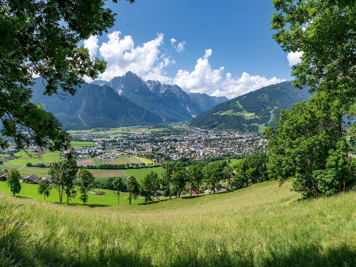 Panorama einer Alpenstadt im Tal, umgeben von majestätischen Bergen und grünen Wiesen unter blauem Himmel mit Wolken.