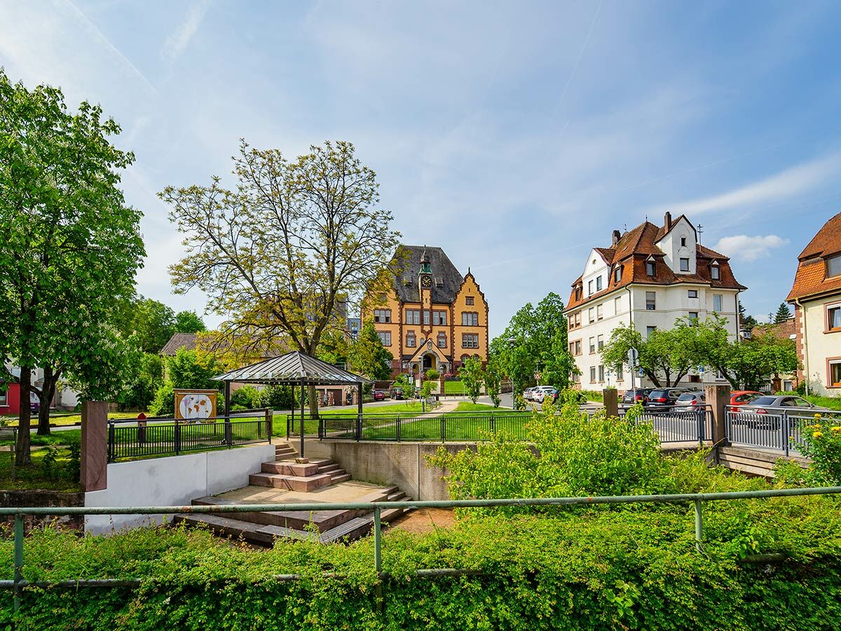 Sonnenbeschienener Stadtpark mit historischem gelbem Gebäude, weißem Wohnhaus, grünem Laub, Pavillon und Weltkarte unter blauem Himmel.