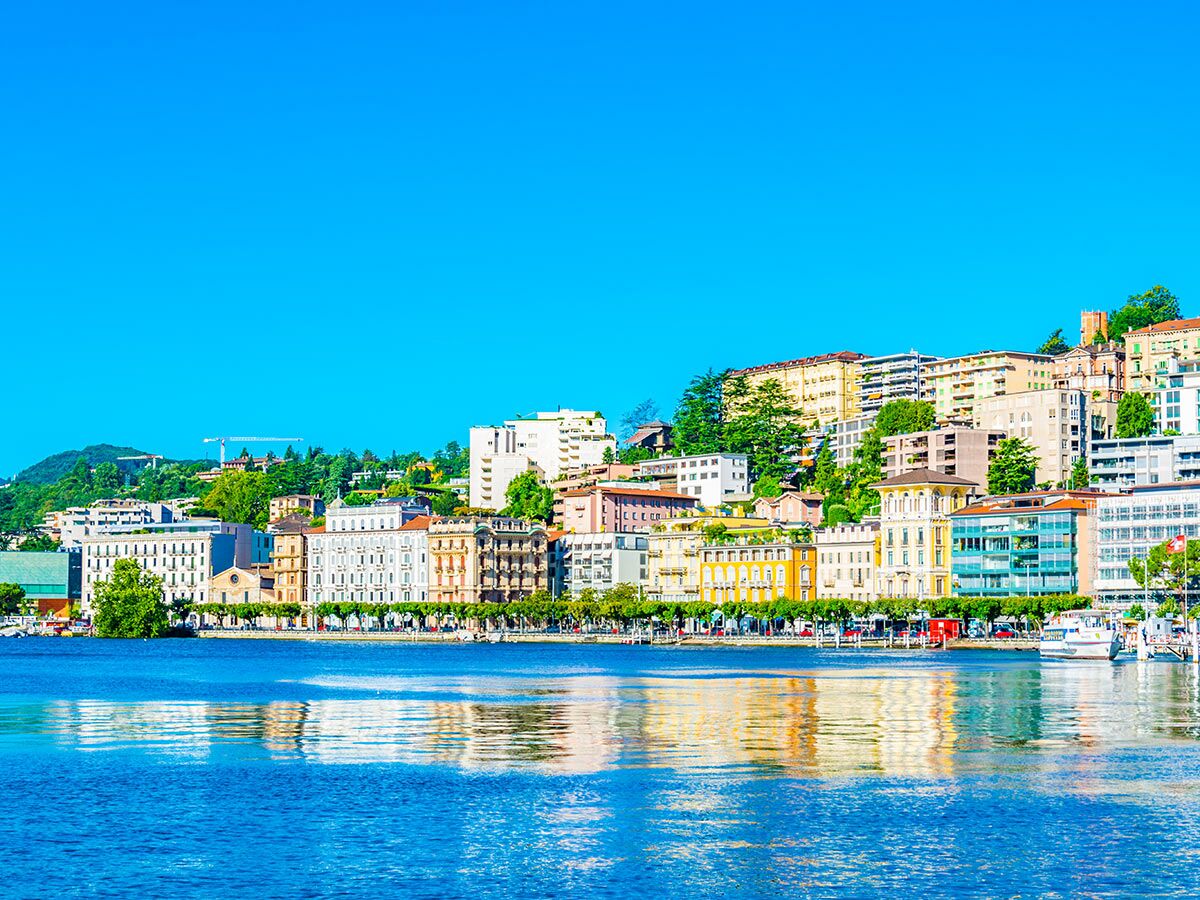 Panoramablick auf die Stadt Lugano am Luganersee mit historischen Gebäuden, grünen Bergen und blauem Himmel.