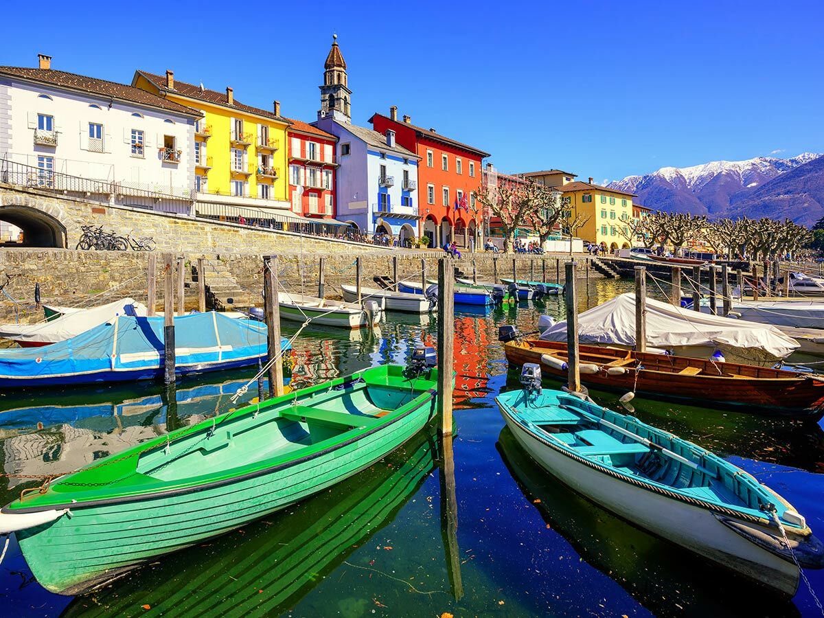 Malerischer Hafen mit bunten Häusern, vielen Booten auf dem Wasser und schneebedeckten Bergen im Hintergrund unter blauem Himmel.