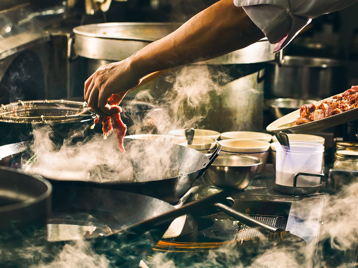 Kochhand fügt rohes Fleischstreifen in einen dampfenden Wok in belebter Küche. Professionelles Kochen mit viel Dampf.
