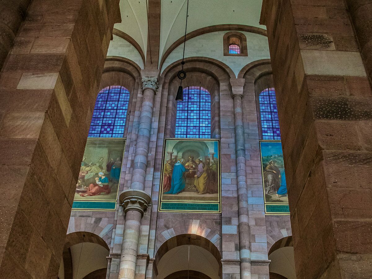 Innenansicht romanische Kathedrale mit massiven Steinsäulen, hohen Bögen, blauen Glasfenstern und drei religiösen Gemälden an der Wand.
