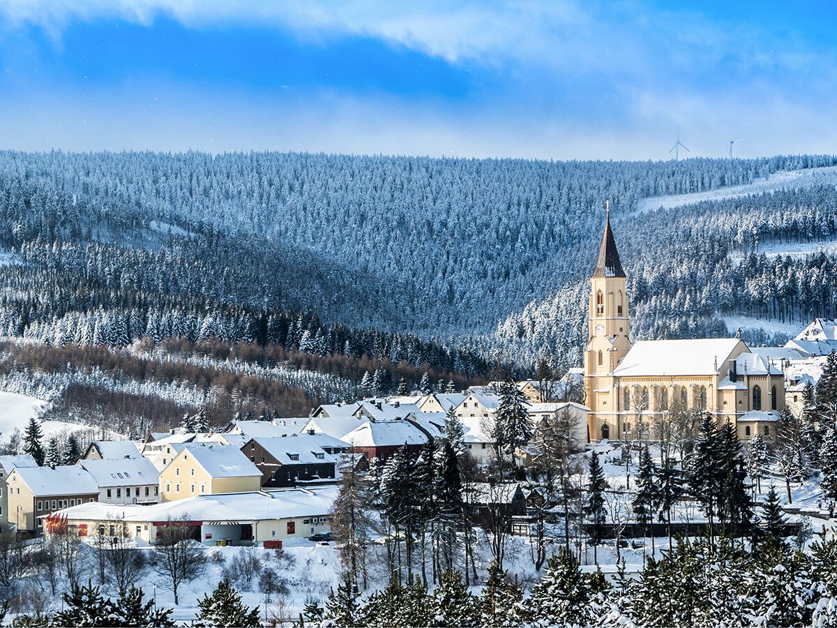 Panoramablick auf ein verschneites Dorf im Winter mit zentraler Kirche, umgeben von schneebedeckten Wäldern und Bergen unter blauem Himmel.