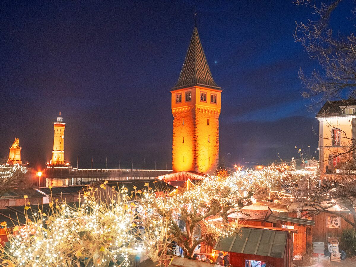 Weihnachtsmarkt Lindau bei Dämmerung, mit hell beleuchtetem Mangturm, Leuchtturm und Löwenstatue am Hafen.