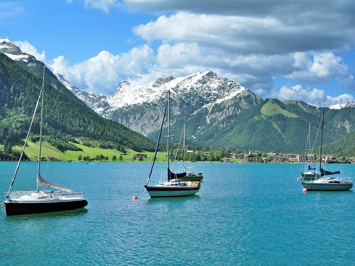 Segelboote auf einem türkisfarbenen Alpensee mit schneebedeckten Bergen, grünen Wäldern und einem Dorf im Hintergrund bei sonnigem Wetter.