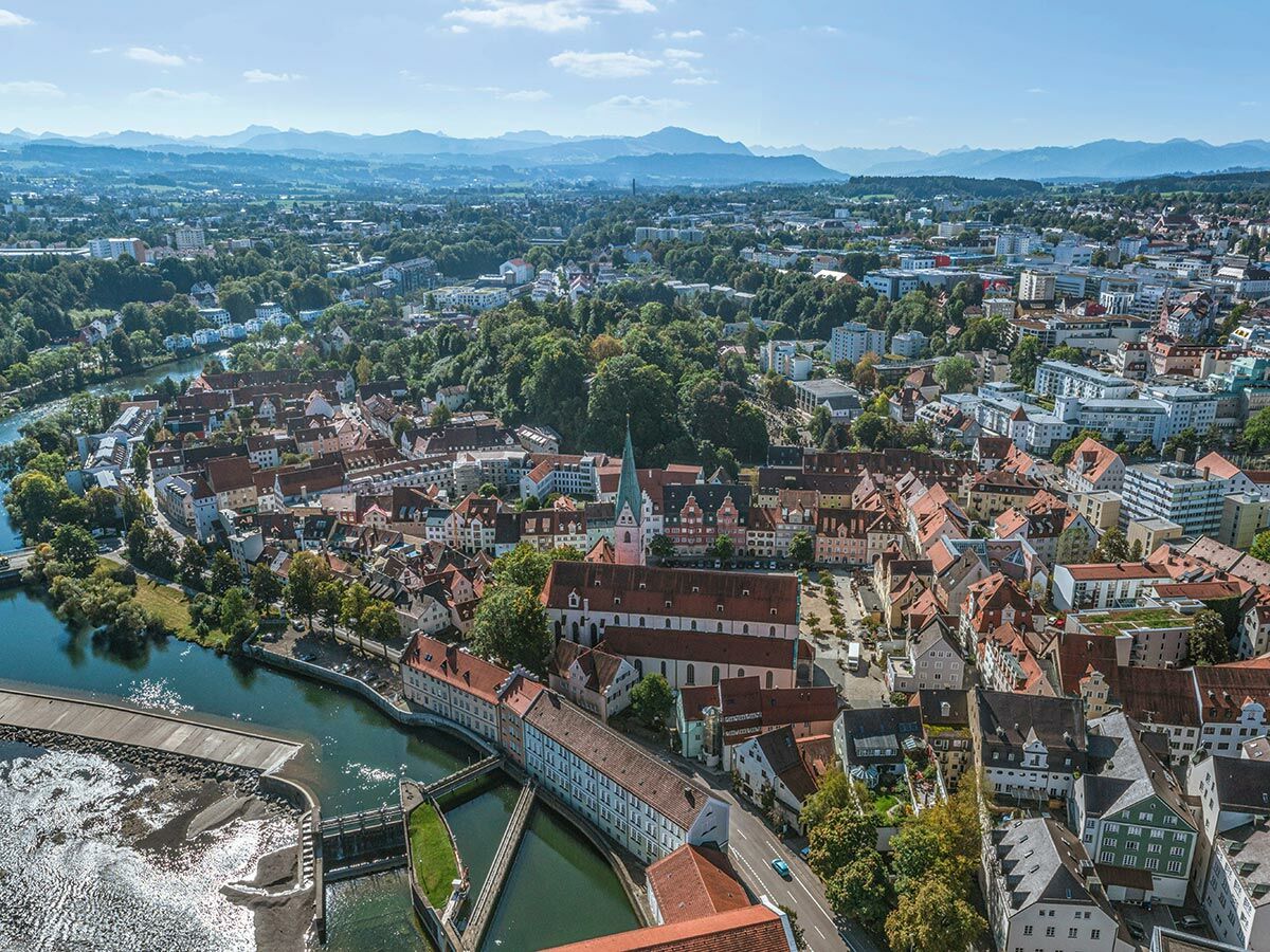 Luftbild der malerischen Altstadt von Füssen am Fluss Lech mit historischen Gebäuden, der Basilika St. Mang und den Alpen im Hintergrund.