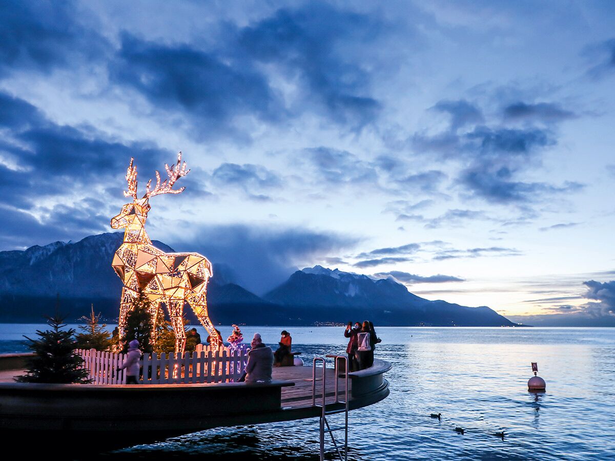 Großes, leuchtendes Rentier auf einem Weihnachtsmarkt am Genfersee mit verschneiten Bergen im Hintergrund bei Dämmerung.