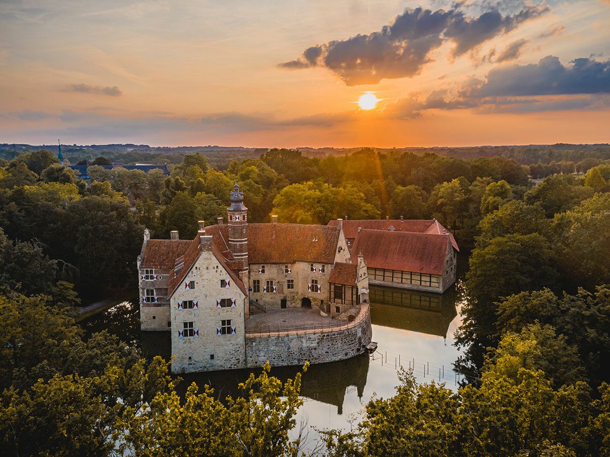 Luftaufnahme eines historischen Wasserschlosses bei Sonnenuntergang, umgeben von dichtem Wald und ruhigem Wasser. Warme Abendsonne.