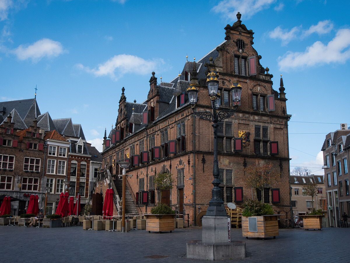 Historisches Rathaus Gouda am Marktplatz mit alten Gebäuden, roten Sonnenschirmen und Laterne bei blauem Himmel.