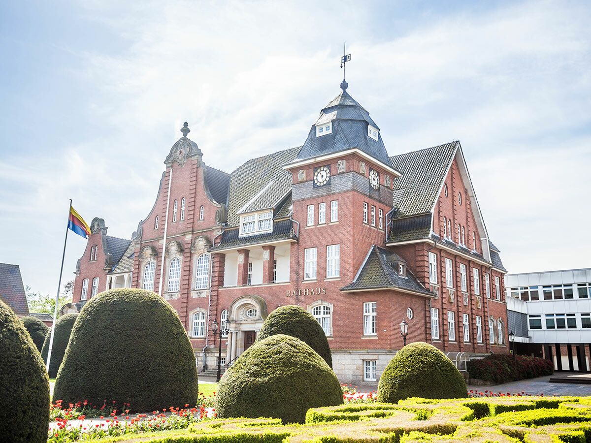 Historisches Backstein-Rathaus mit Uhrturm und Flagge, umgeben von einem gepflegten Garten mit Formschnitt-Bäumen und roten Blumen.
