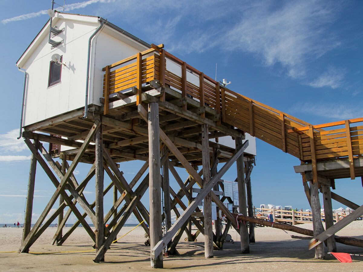 Weißer Pfahlbau mit Holzstegen auf sandigem Strand unter blauem Himmel. Im Hintergrund sind Strandbesucher und Strandkörbe zu sehen.