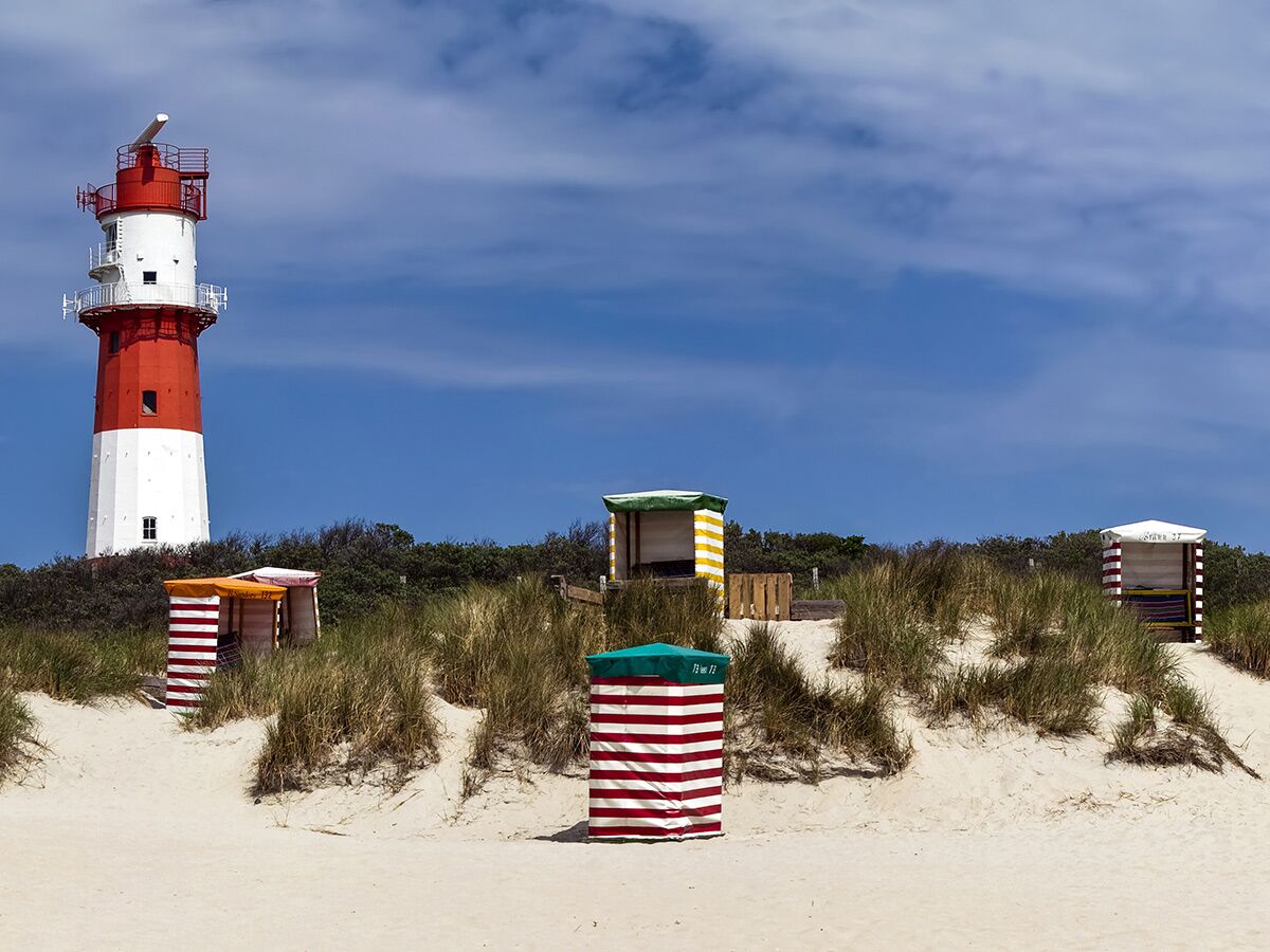 Roter und weißer Leuchtturm am Sandstrand mit Dünen und bunten Strandkörben unter blauem Himmel.