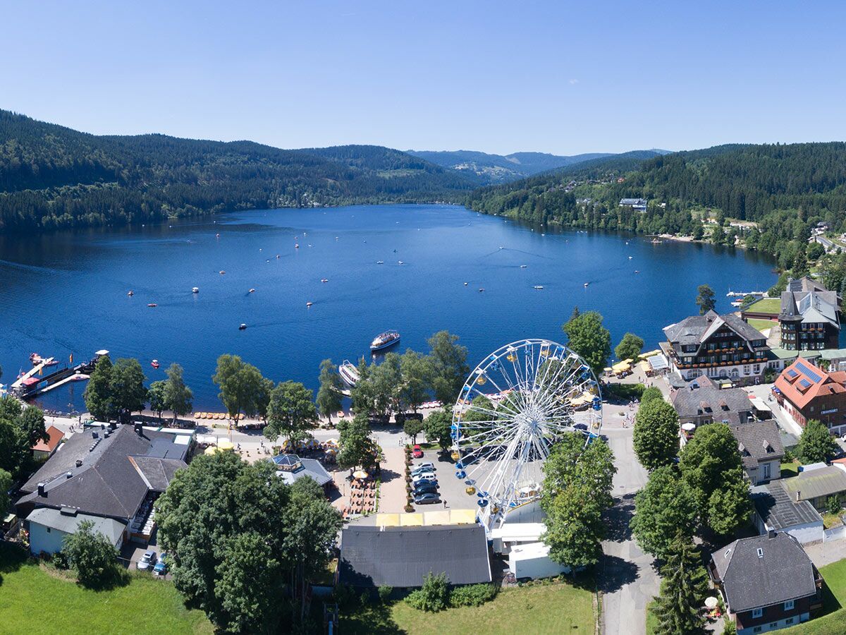 Luftbild des Titisees im Schwarzwald mit Riesenrad am Ufer, Häusern, Wald und vielen Booten auf dem See.