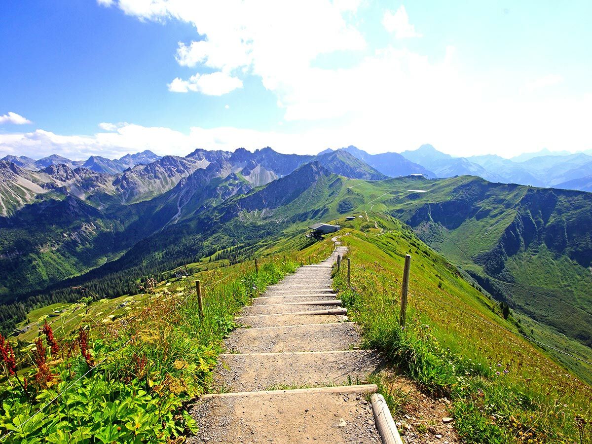 Wanderweg mit Stufen auf einem grünen Bergrücken im Sommer. Weites Alpenpanorama mit Tälern und fernen Gipfeln unter blauem Himmel.