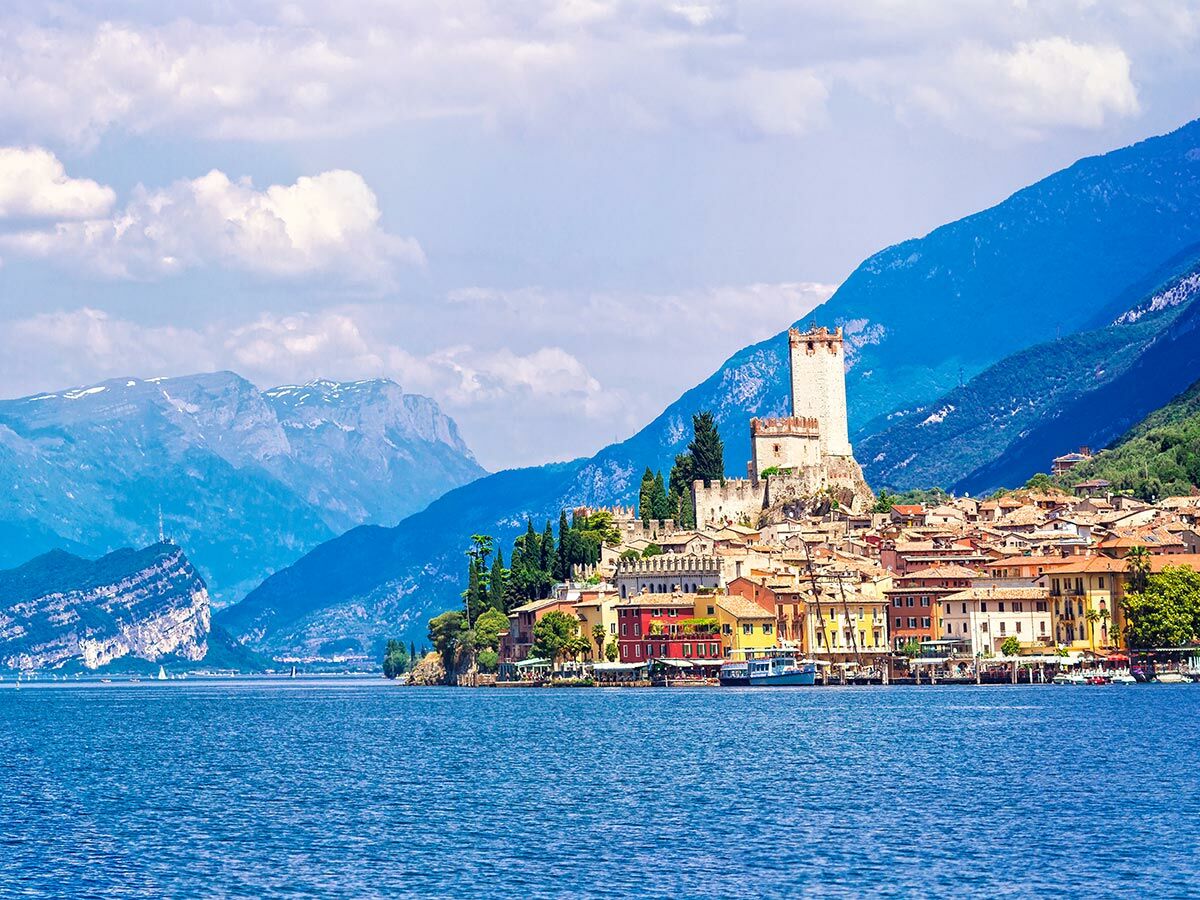 Blick auf Malcesine am Gardasee mit Scaligerburg, bunten Häusern, Bergen und Booten auf blauem Wasser unter Wolkenhimmel.