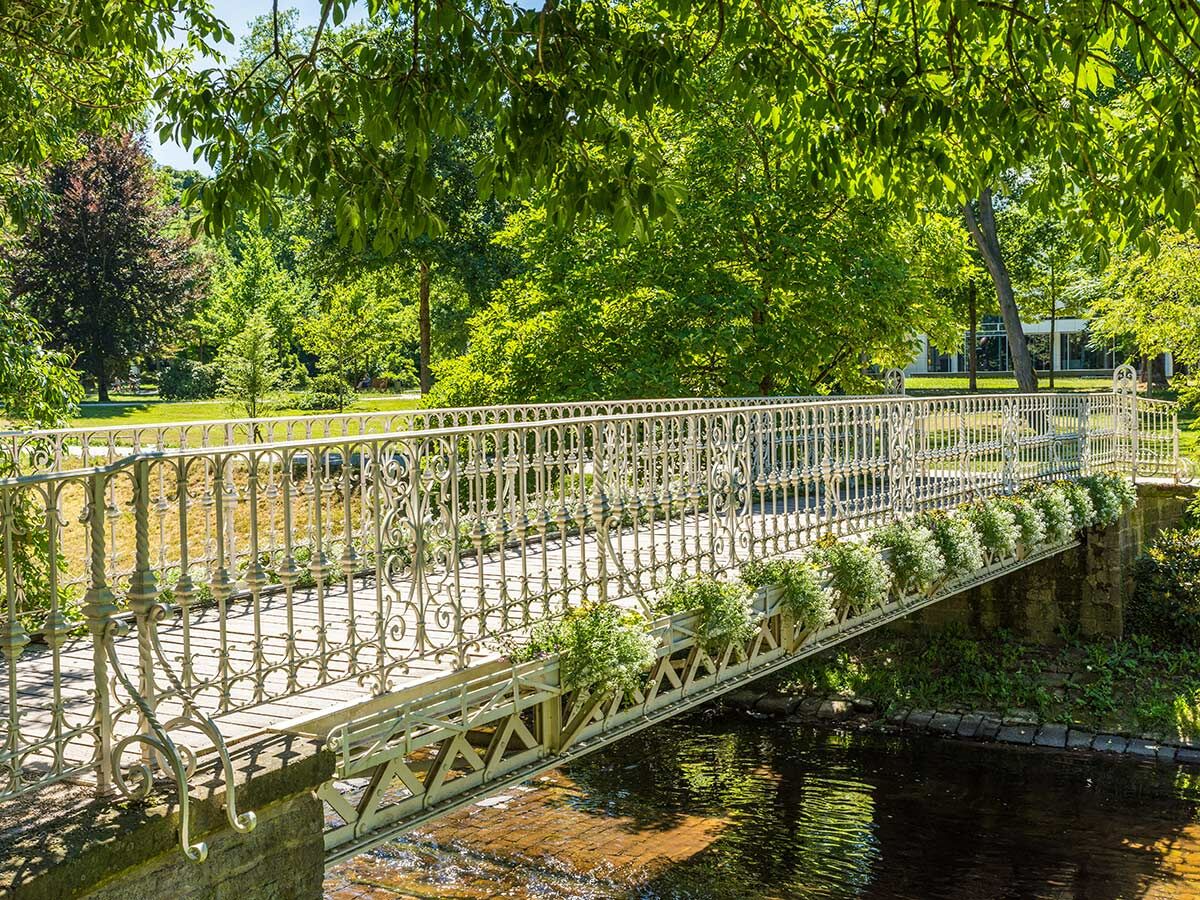 Blick auf eine weiße, verzierte Fußgängerbrücke über einen Bach in einem üppig grünen Park an einem sonnigen Tag. Blumenkästen schmücken das Geländer.