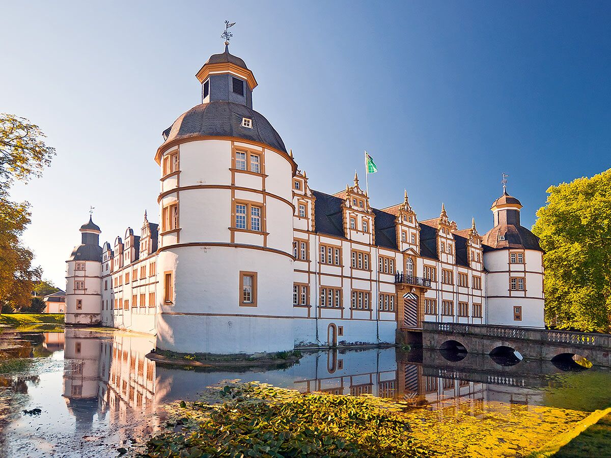 Historisches Wasserschloss Raesfeld mit weißen Fassaden und braunen Details, gespiegelt im Moat unter blauem Himmel.