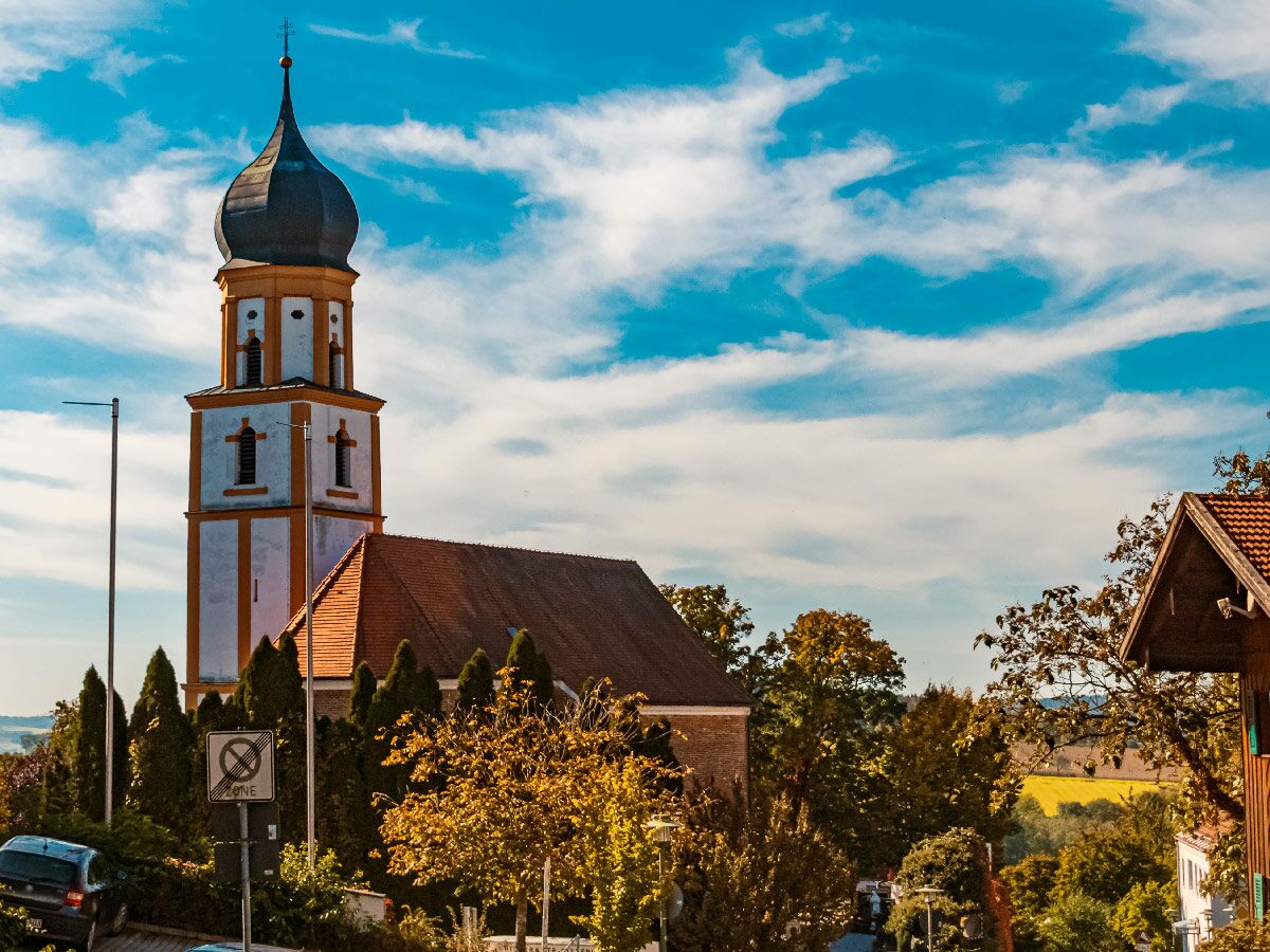 Hoher Kirchturm mit Zwiebelhaube und rotem Dach des Kirchenschiffs unter blauem Himmel mit weißen Wolken, umgeben von Bäumen und Gebäuden.