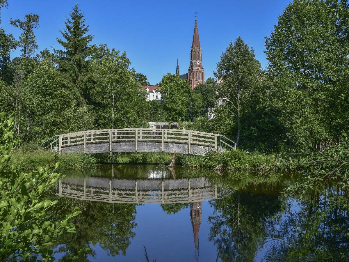 Hölzerne Brücke über ruhigem Gewässer, umgeben von grünen Bäumen, mit klarer Spiegelung eines roten Kirchturms und Brücke.