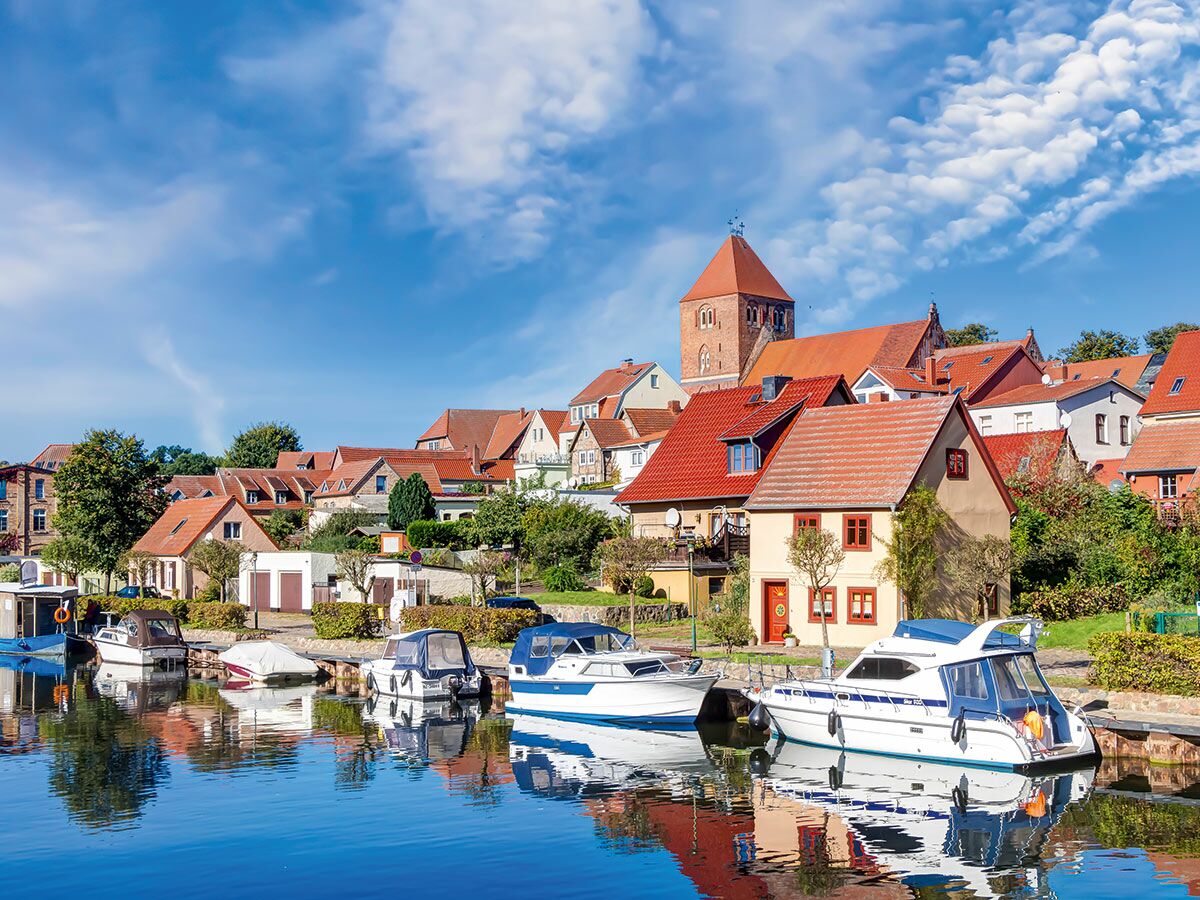 Idyllisches Küstendorf mit roten Dächern, Kirchturm und im Wasser spiegelnden Booten unter blauem Himmel.