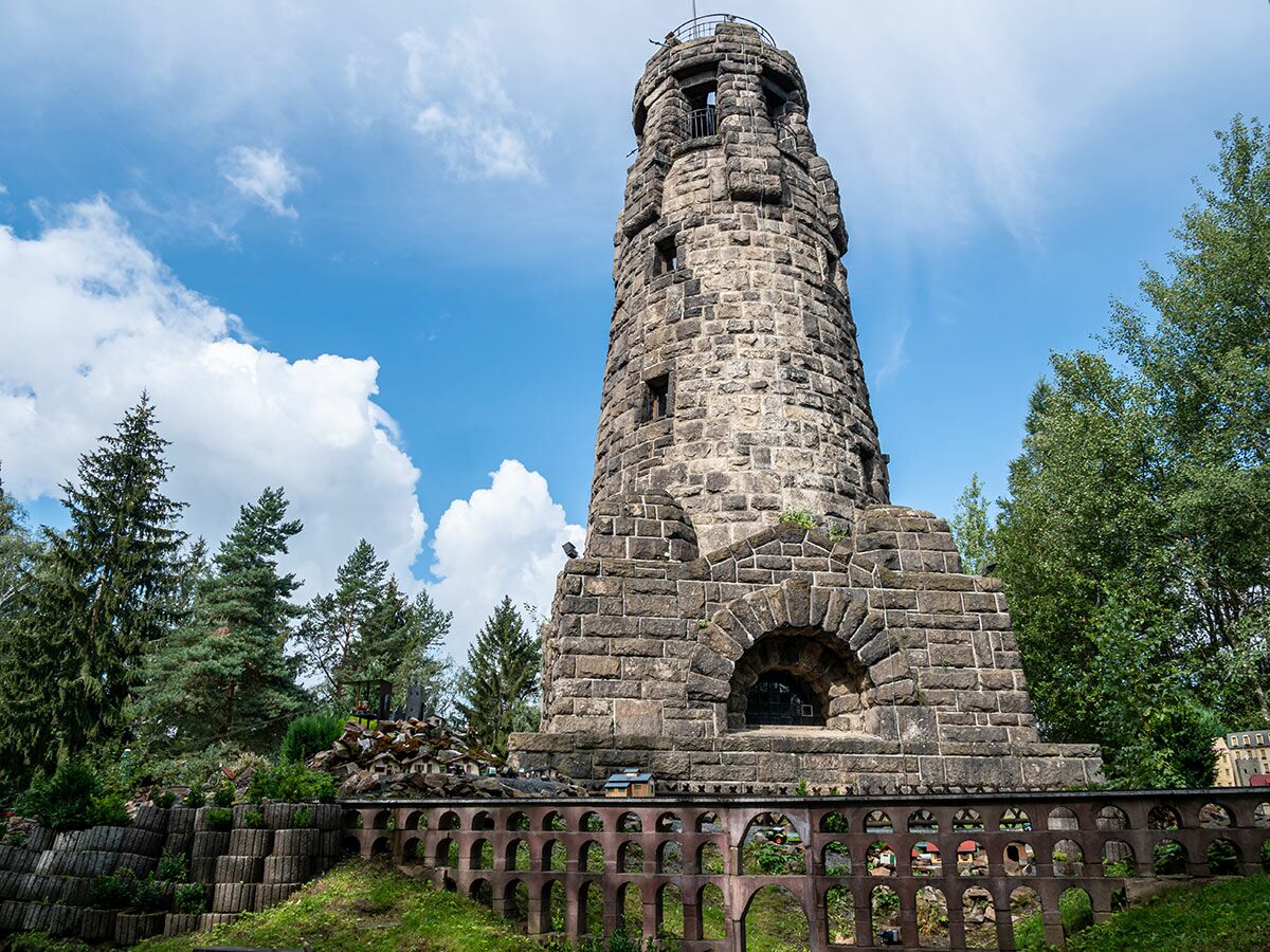 Historischer Bismarckturm aus Stein in Viersen mit blauem Himmel, Wolken und Bäumen umgeben von einer Miniaturlandschaft.