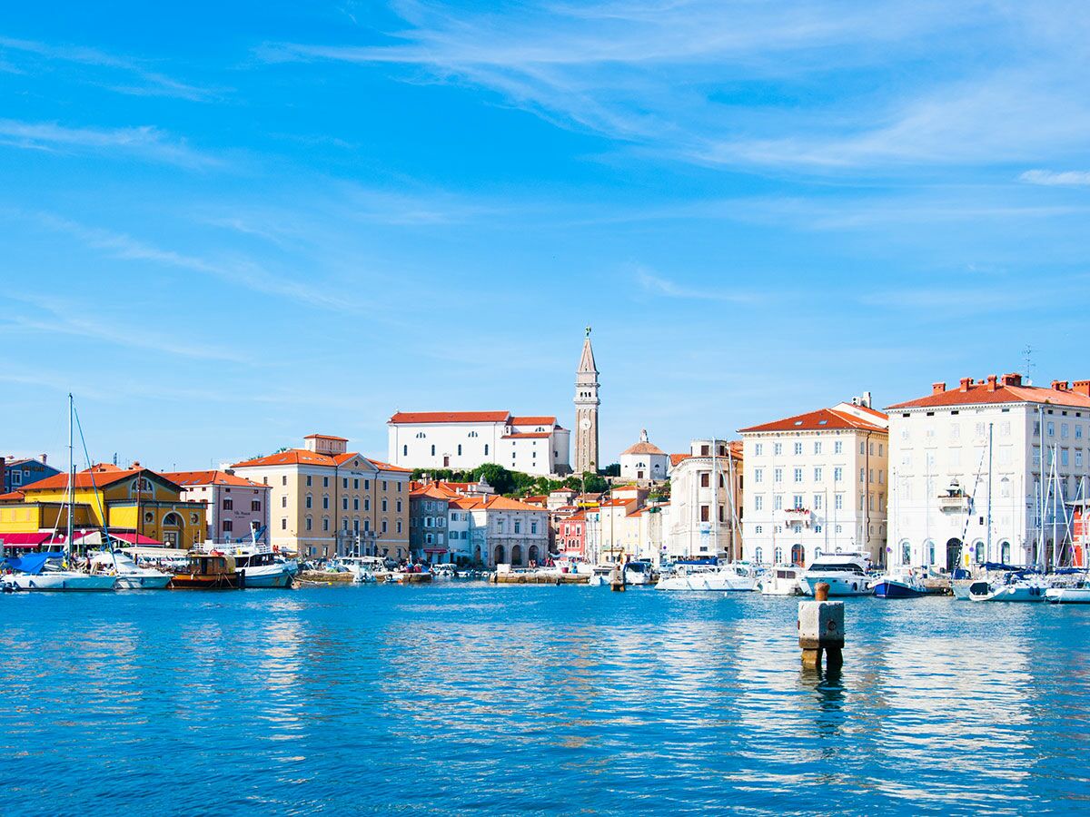 Panoramablick auf die slowenische Hafenstadt Piran mit bunten Gebäuden, Segelbooten, Yachten und dem Glockenturm unter blauem Himmel.