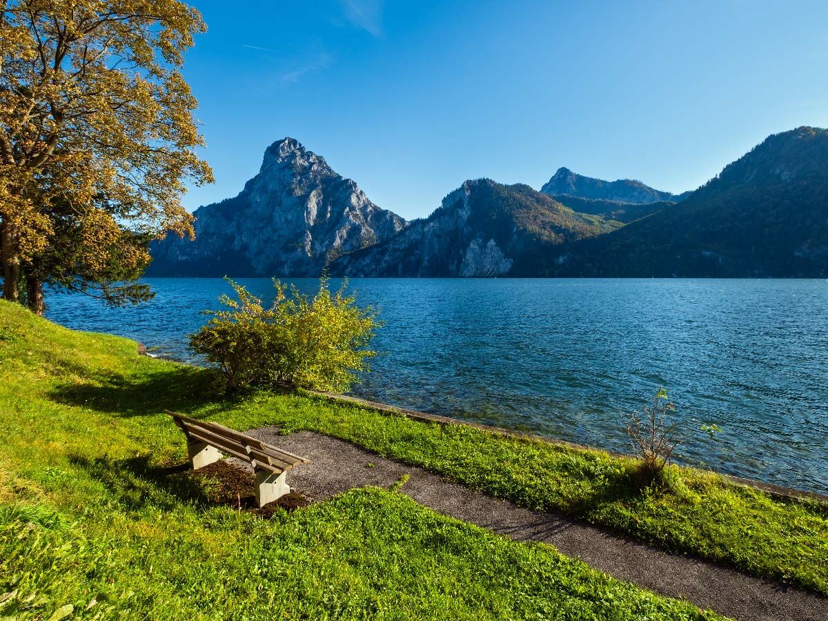 Idyllischer Bergsee mit Felsbergen, herbstlichem Baum, Holzbank und Uferweg unter klarem Himmel in den Alpen, Österreich.