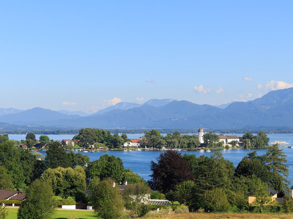 Blick auf den Starnberger See mit einer Insel, einem Kloster, Häusern und grünen Bäumen. Im Hintergrund erheben sich blaue Alpenberge unter klarem Himmel.