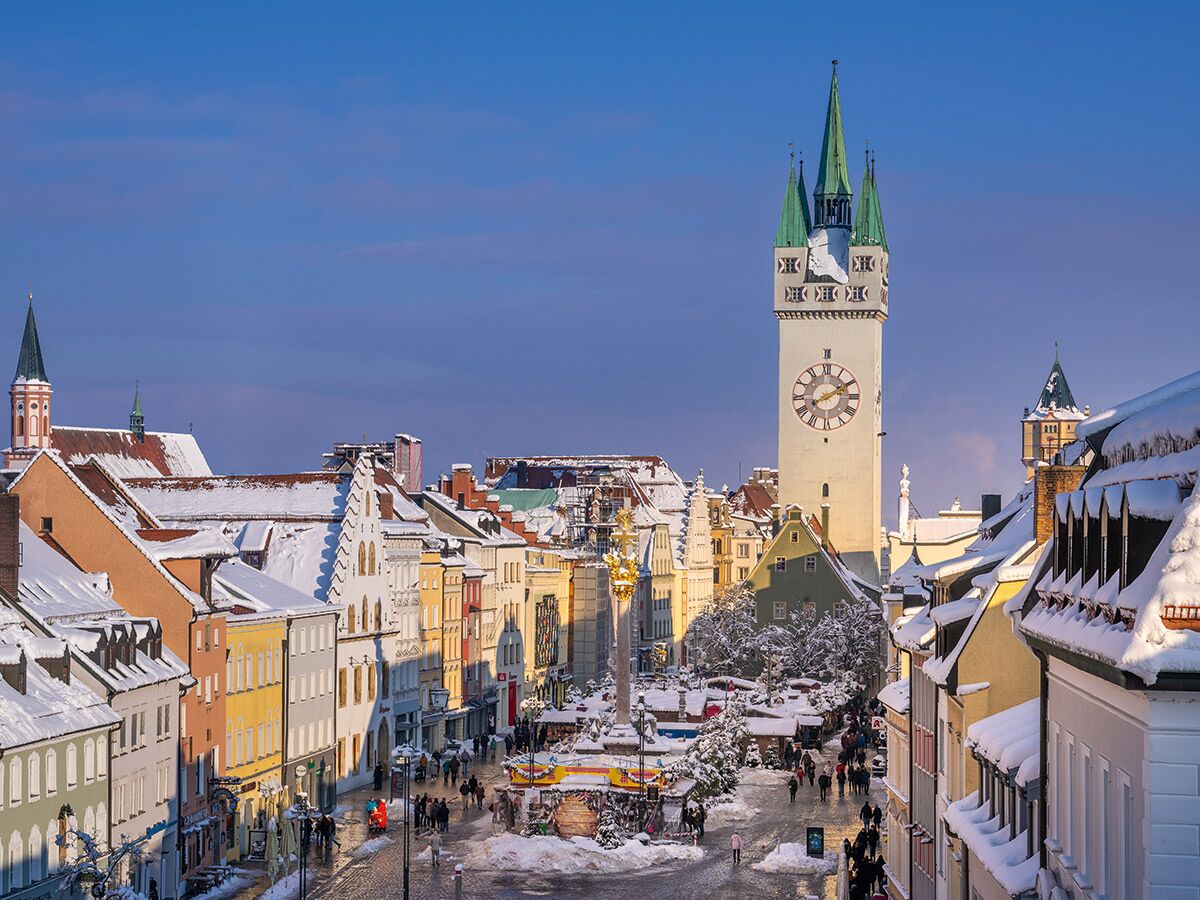 Blick über den schneebedeckten Stadtplatz von Straubing in Bayern mit historischen Häusern, belebten Gassen und dem markanten Stadtturm unter blauem Himmel.