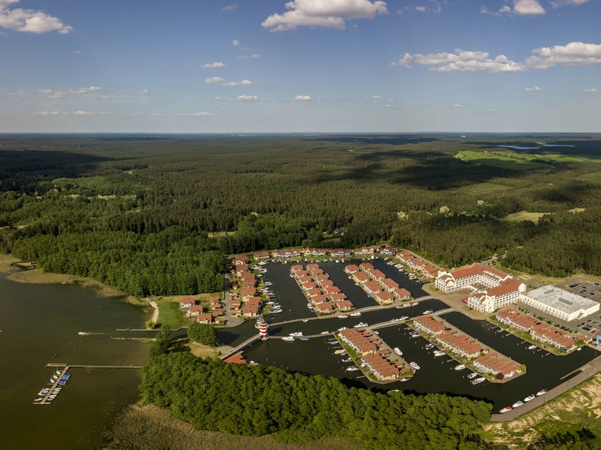 Luftbild eines Marina-Feriendorfes mit vielen Häusern, Booten, Hotel und Leuchtturm am Waldsee unter blauem Himmel.