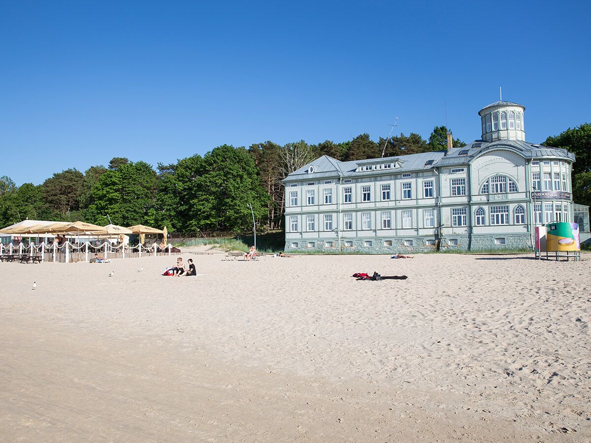Sandstrand Jūrmala mit historischem Holzgebäude, Café, entspannten Menschen unter blauem Himmel und Bäumen.
