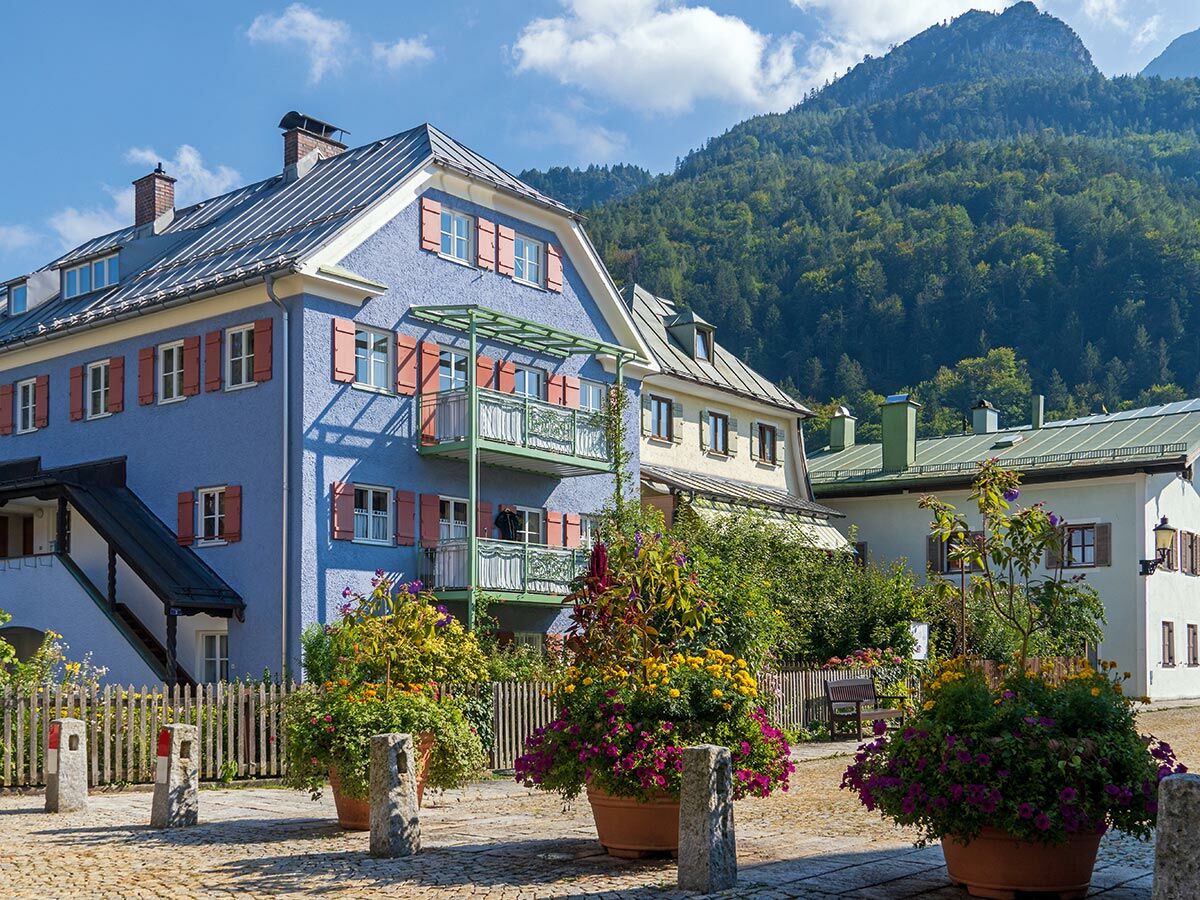 Blaues Haus mit roten Fensterläden und Balkonen in einem malerischen Bergdorf, umgeben von bunten Blumen und grünen Bergen.