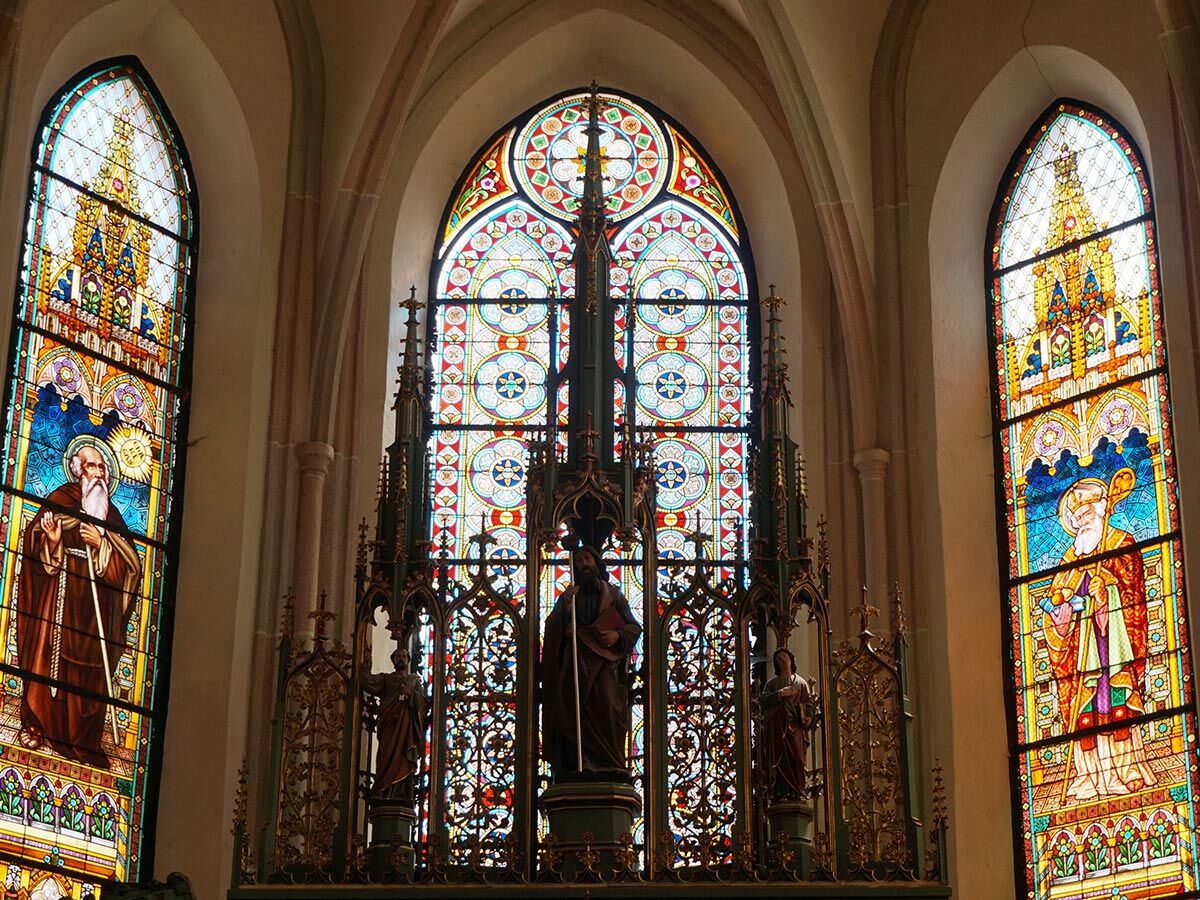 Drei farbenprächtige gotische Buntglasfenster in einer Kirche, darunter die Darstellungen von Heiligen und ein reich verzierter Altaraufsatz mit Statuen.