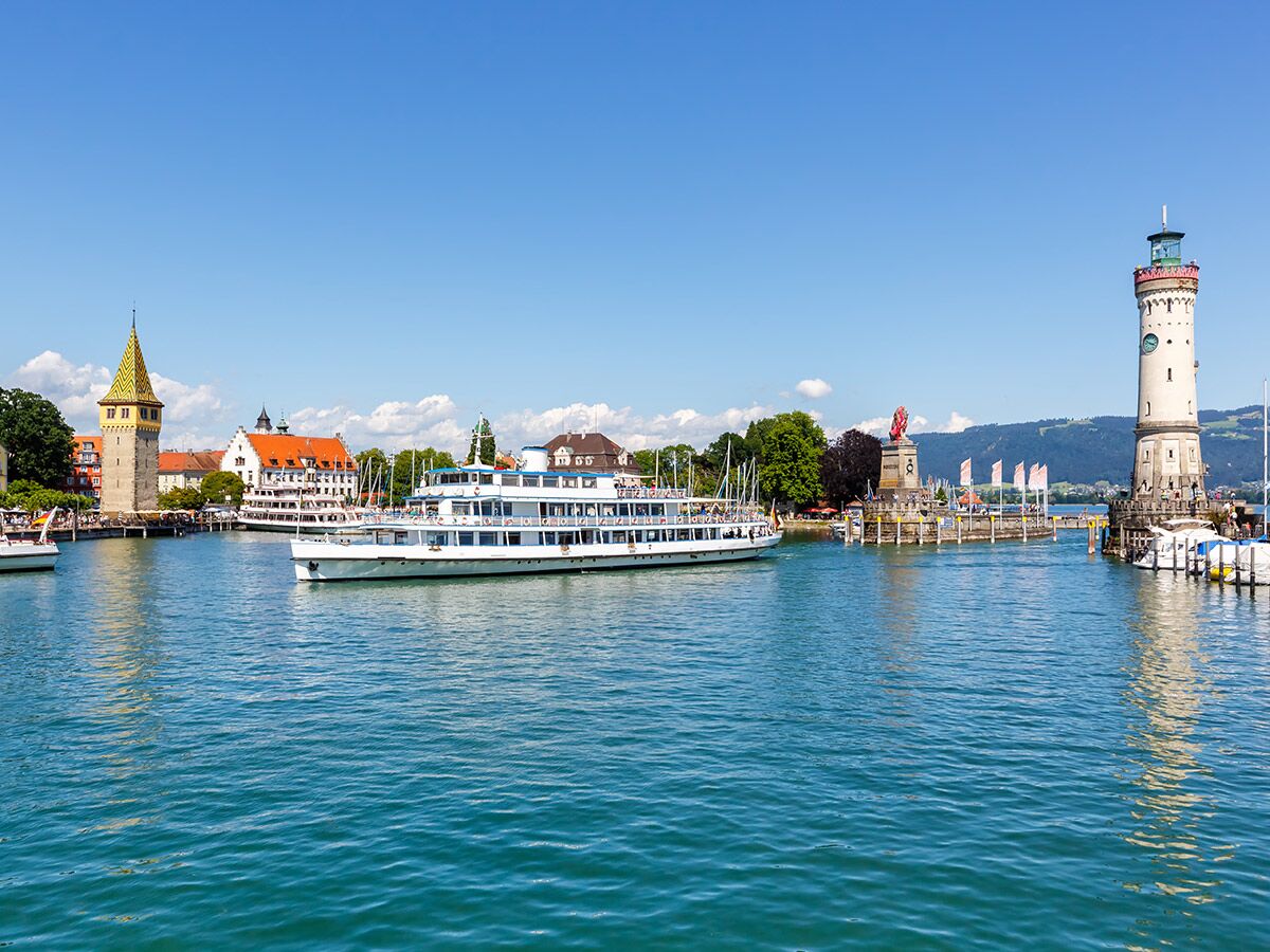 Panoramablick auf den Lindauer Hafen am Bodensee mit weißem Leuchtturm, Mangturm, Löwenstatue und Schiffen bei Sonnenschein.