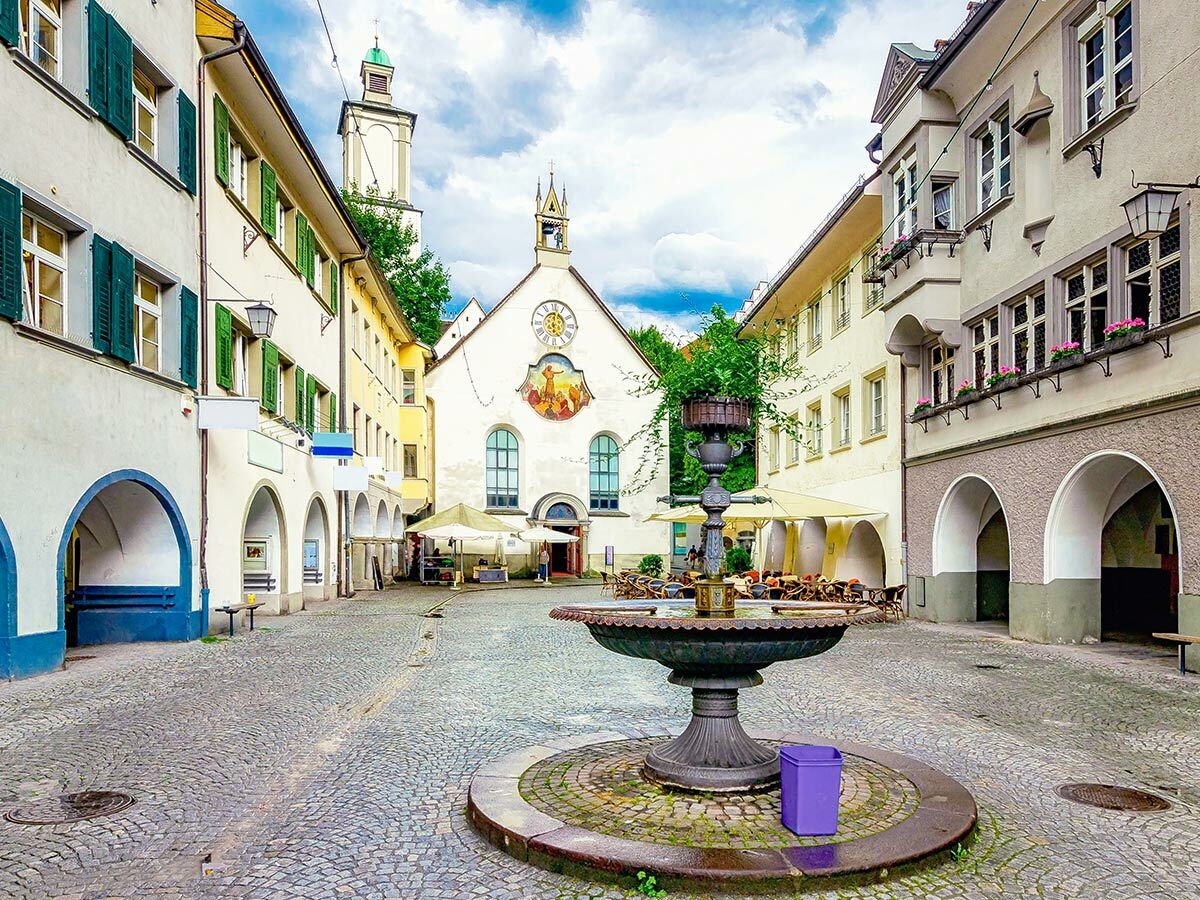 Blick auf einen malerischen historischen Stadtplatz in Europa mit altem Brunnen, Kirche, Gebäuden und Außencafés.
