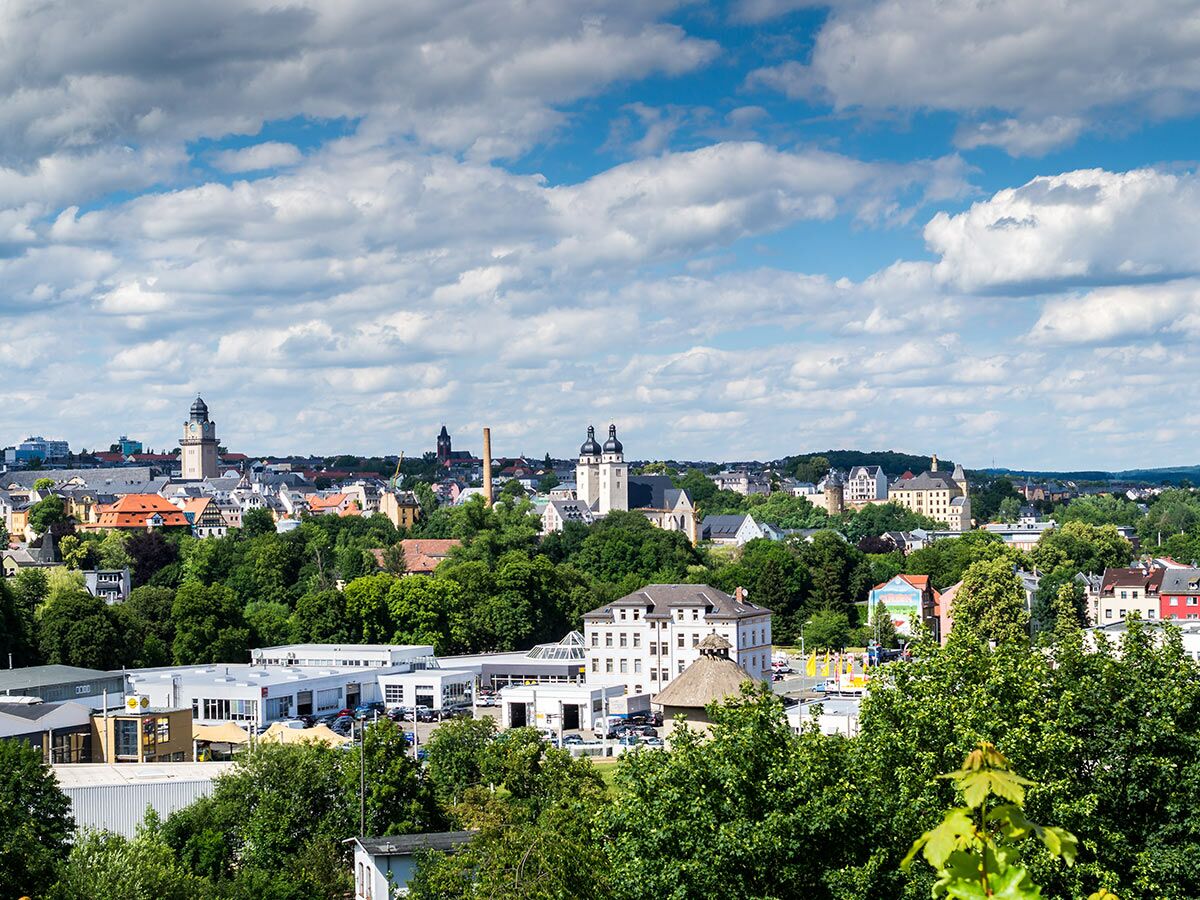 Panoramablick auf eine europäische Stadt mit historischen Gebäuden, Kirchen, bewaldeten Hügeln und blauem Himmel mit weißen Wolken.