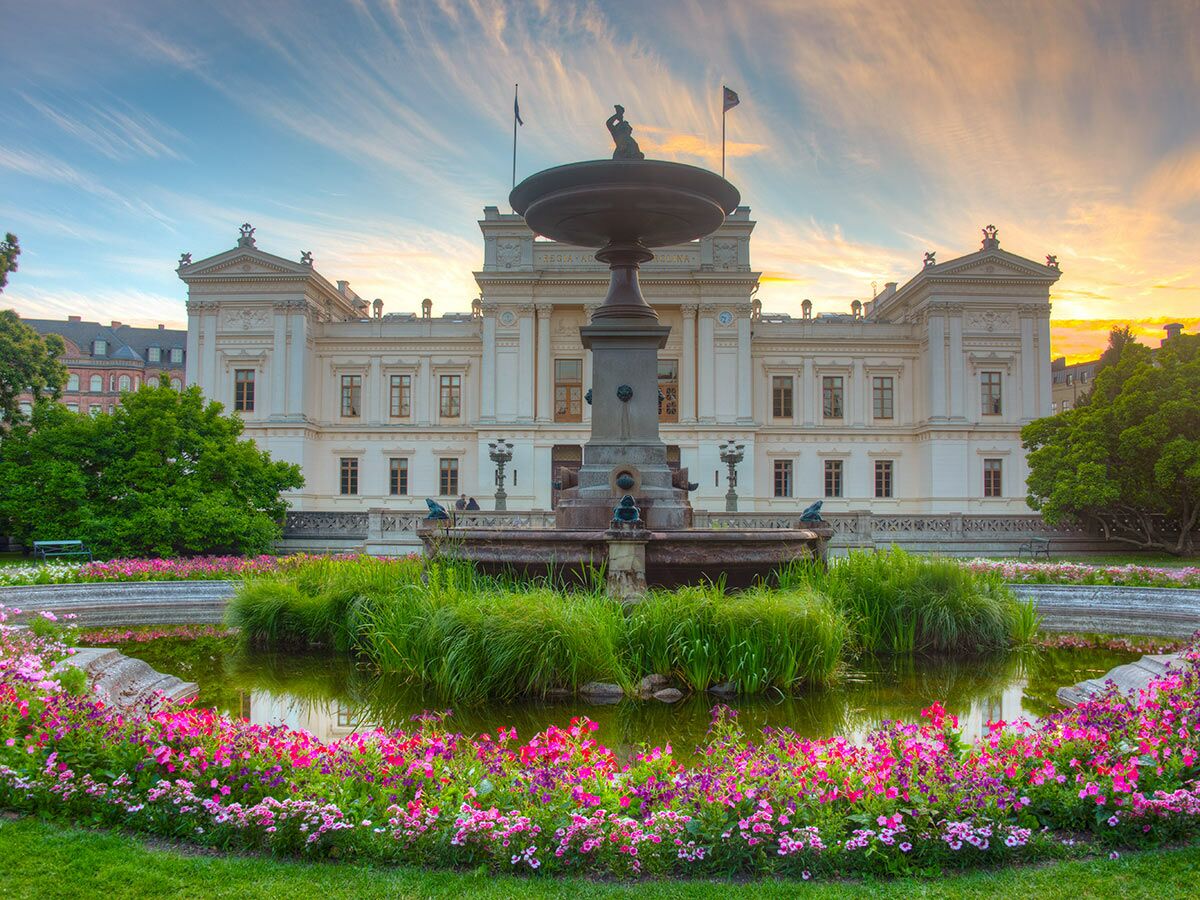 Das historische Universitetshuset in Lund bei Sonnenuntergang, umgeben von einem Brunnen, Teich und rosa Blumen.