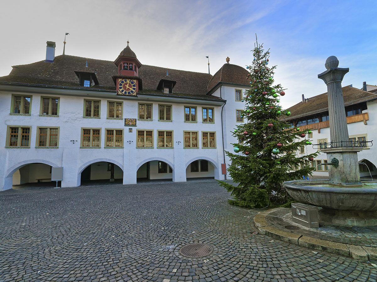 Historischer Rathausplatz mit weißem Gebäude, Uhrturm, Arkaden, Kopfsteinpflaster, geschmücktem Weihnachtsbaum und Steinbrunnen.