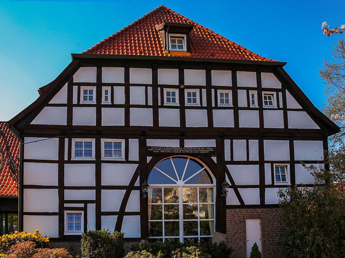 Historisches Fachwerkhaus mit weißer Fassade, dunklen Holzbalken und rotem Ziegeldach unter blauem Himmel.