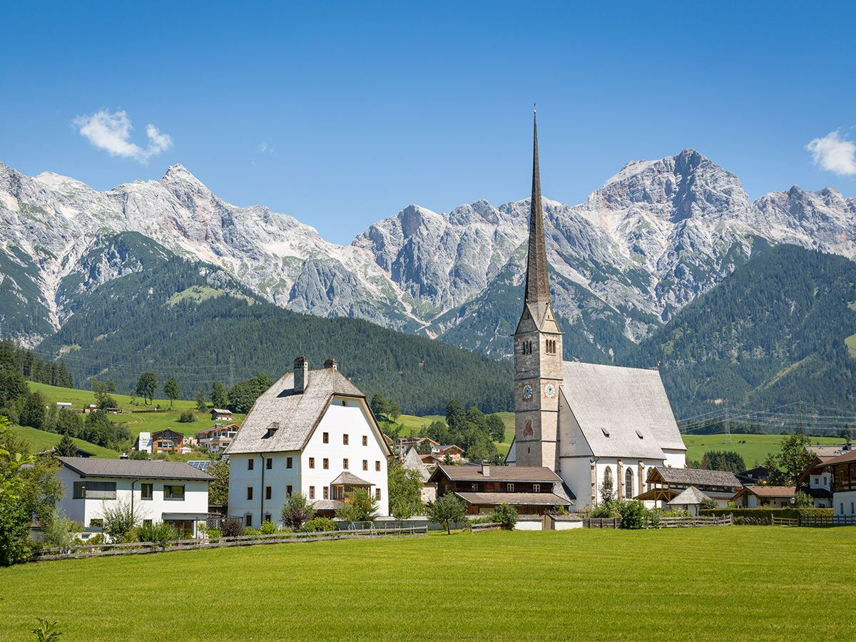 Malerisches Bergdorf mit Kirche, grünen Wiesen, traditionellen Häusern und majestätischen Alpen unter blauem Himmel.