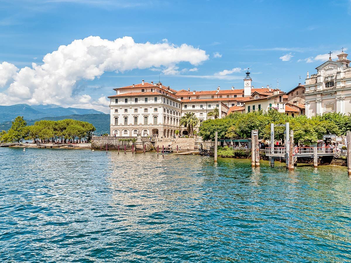 Blick über den Lago Maggiore auf Isola Bella mit dem Borromeo-Palast, einer Kirche und Anlegestellen. Berge im Hintergrund.