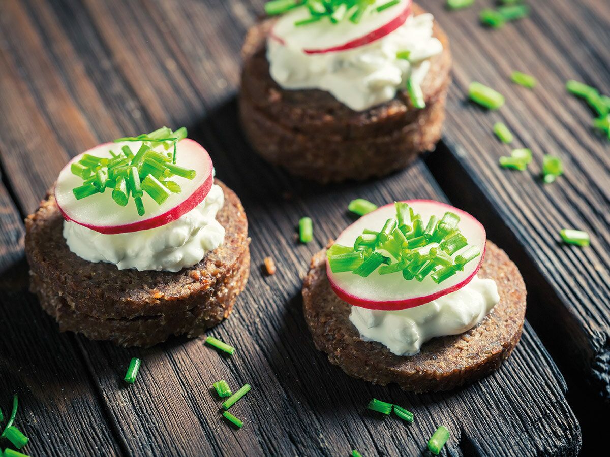 Kleine Roggenbrot-Canapés mit Frischkäse, Radieschenscheiben und Schnittlauch auf rustikalem Holzbrett.