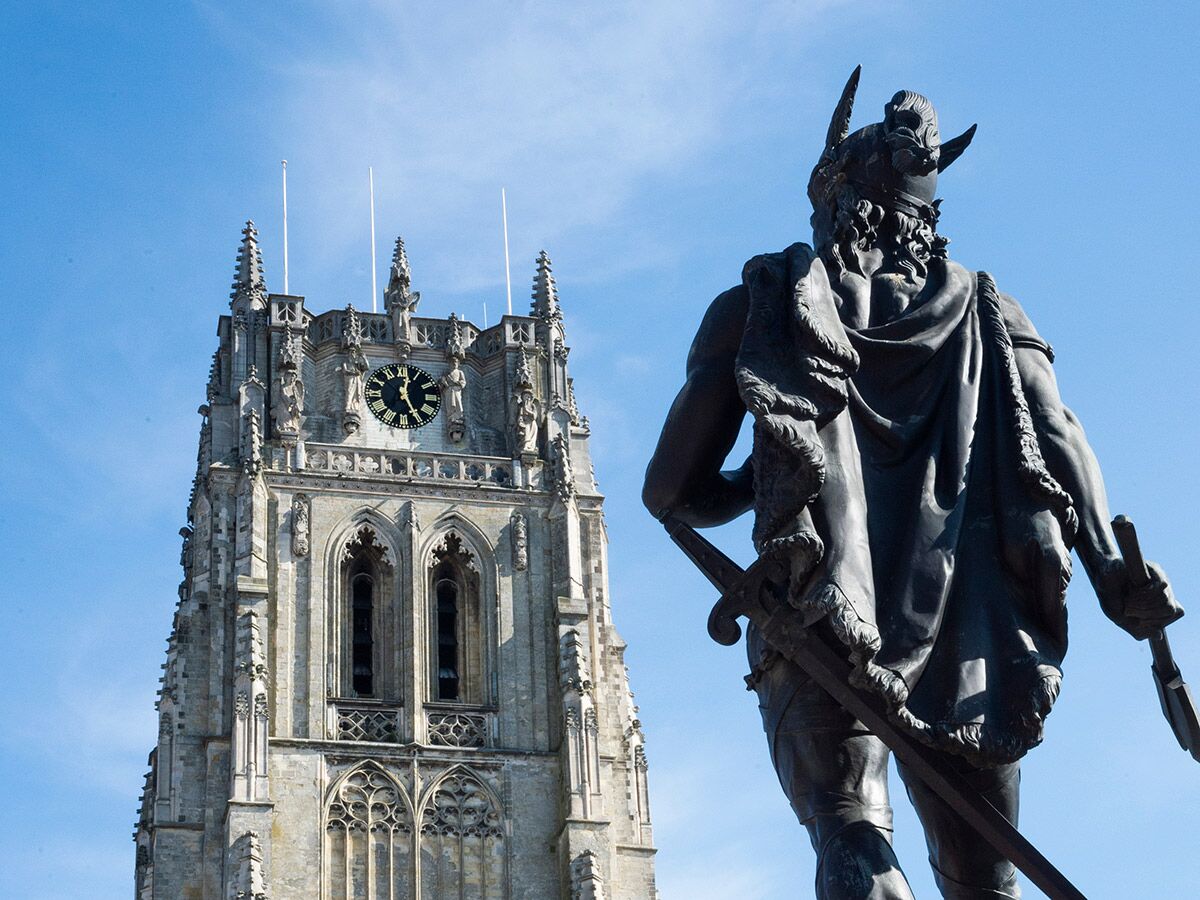 Statue eines Kriegers mit Schwert vor einem gotischen Kirchturm mit Uhr unter blauem Himmel.