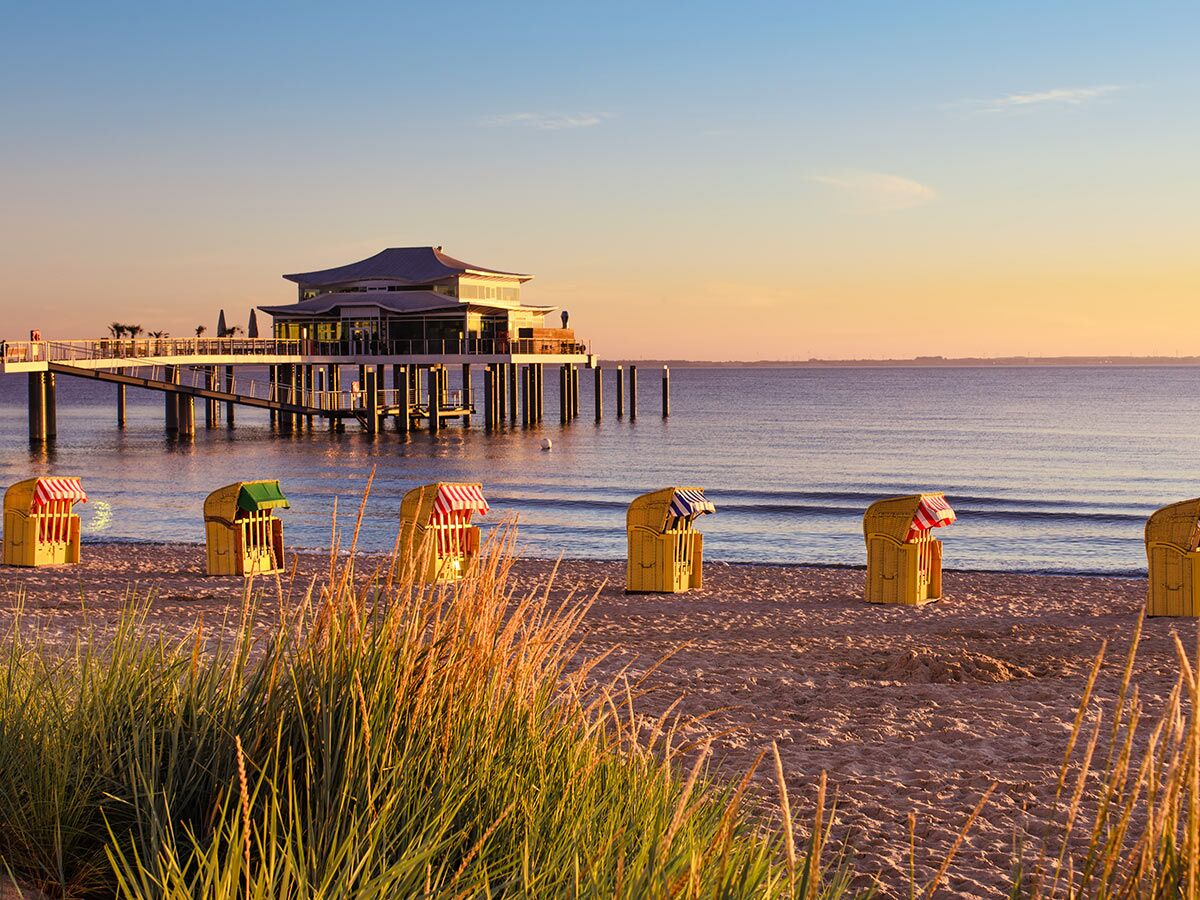 Idyllischer Strand mit traditionellen Strandkörben, einem langen Pier und Gebäude bei Sonnenuntergang an der Ostsee.