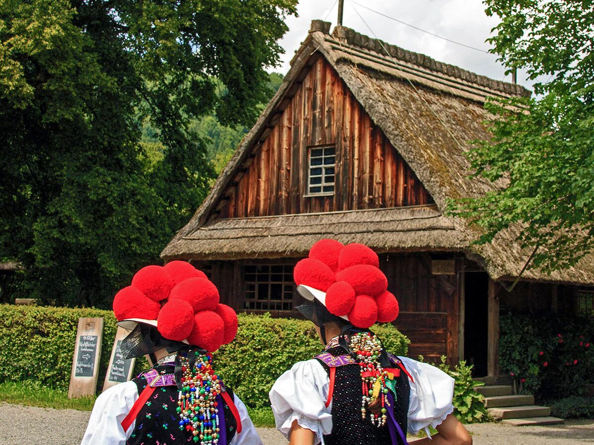 Zwei Personen mit rotem Bollenhut und Schwarzwälder Tracht vor einem Reetdach-Bauernhaus im Grünen.