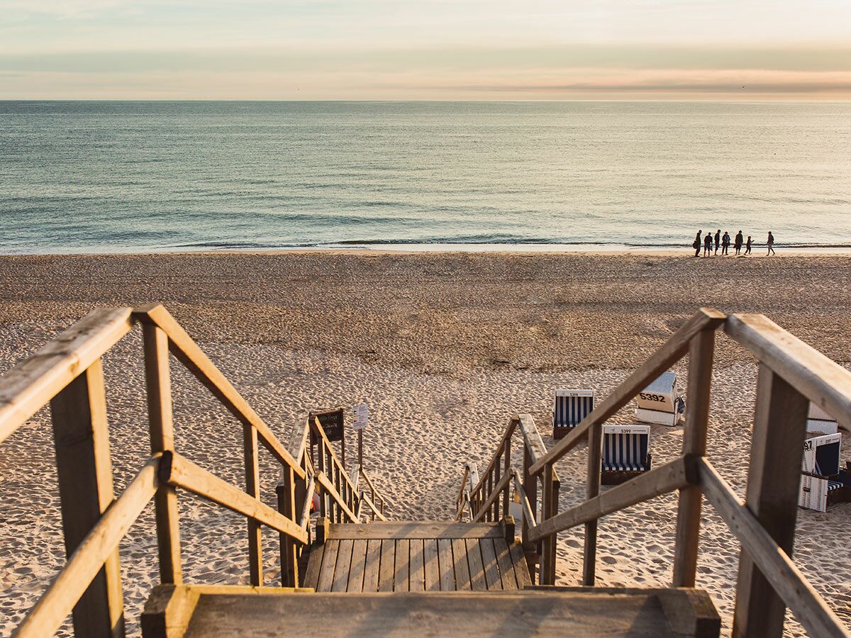 Hölzerne Treppe führt zu einem sandigen Strand mit Strandkörben und dem Meer, Menschen gehen am Wasser entlang.
