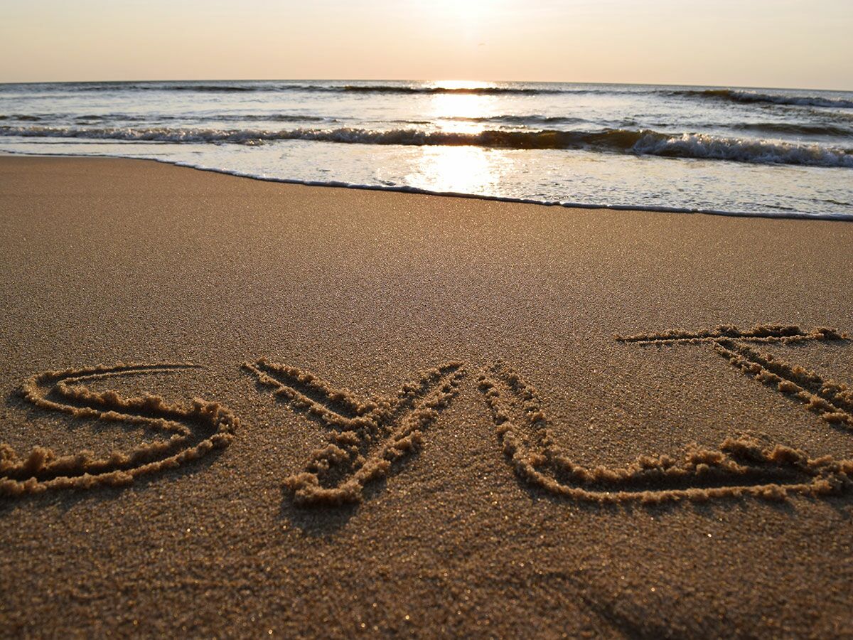 Der Schriftzug 'SYLT' ist in den goldenen Sand eines Strandes geschrieben, mit Wellen und einem warmen Sonnenuntergang im Hintergrund.