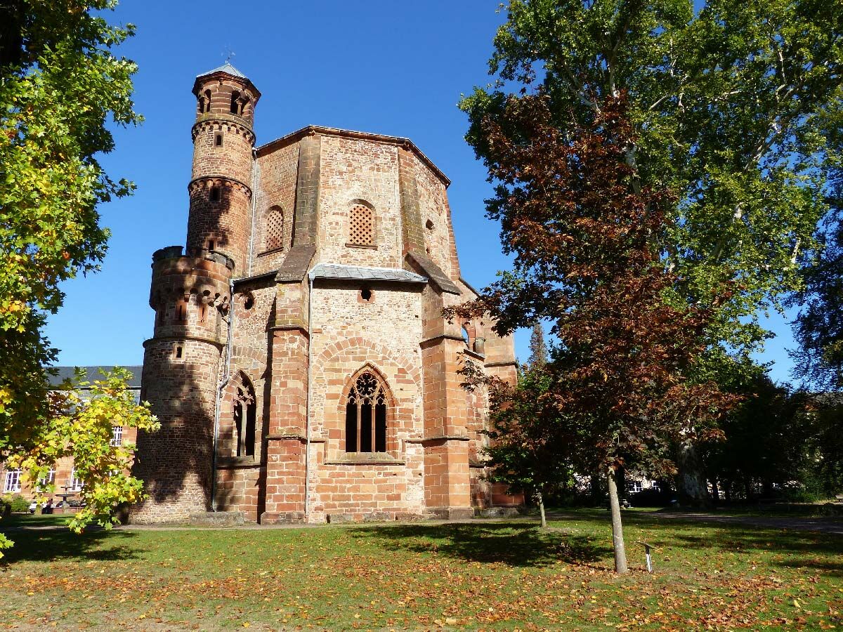 Historische Königshalle Lorsch im Herbst, roter Sandsteinbau, Rundtürme, bunte Bäume unter blauem Himmel.