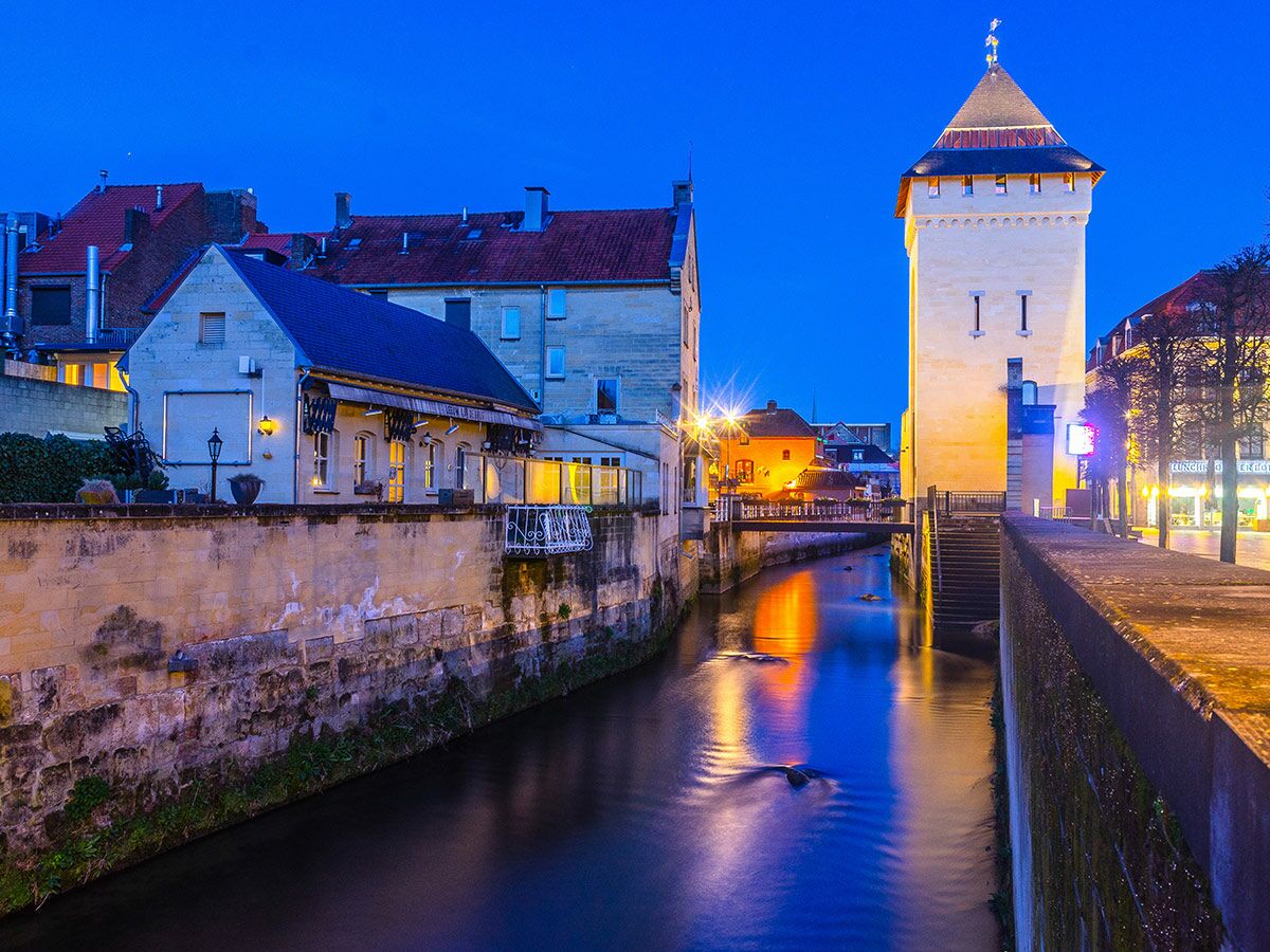 Abendaufnahme eines Stadtkanals mit historischen Gebäuden, markantem Turm und Lichtreflexionen im Wasser.